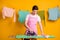 Photo of young happy handsome cheerful positive focused man ironing washed clothes isolated on yellow color background