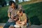 Photo of young man and boy with phones in hands sitting behind wooden fence in park