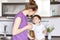 Photo of young woman in casual clothing stands against kitchen interior, looks at her adorable son with great love, gives tasty dr