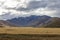 Photograph of a mountain range with grey clouds located behind a large brown agricultural field in New Zealand