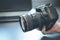 Photographer holds a reflex camera with telephoto lens in his hand. Table and laptop in the blurry background