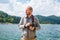 Photographer man in hoodie walking around lake with DSLR camera and shooting nature, half body portrait