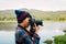 Photographer in wearing plaid shirt and blue wool hat using Camera DSLR Digital Single Lens Reflex take a photo river and Mountain