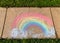 The phrase `Be Happy` written with sidewalk chalk, inside a rainbow, on gray concrete pavement background