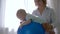 Physiotherapist trains male baby on the big blue ball near window in room closeup