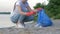 Picking garbage, volunteer woman in rubber gloves collects plastic trash in refuse bag while cleaning coast close up