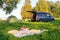 Picnic date in forest, tent and camp chairs, gray minibus with open door, blanket, wicker basket, wine glasses, snacks, fruit,