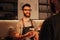 A picture of bearded hipster barmen that wears glasses standing behind the bar stand and holding a cup of coffe that he