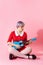 Picture of beautiful dollish girl with short light violet hair wearing red dress playing blue ukulele over pink