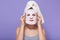 Picture of caring cute young female applying face mask, moisturizing skin, caring about herself, having towel on head, standing