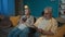In the picture the elderly couple sit on the sofa in the apartment against the blue wall. The man reads the book aloud