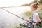 A picture of girl sitting alone at river shore. She is fishing. Girl is holding fish-rod with both hands. She is looking