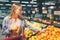 A picture of a girl standing near big basket of citrus. She has chosen one orage and trying to choose some more of it