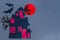 A picture made of paper on the theme of Halloween. A black witch on a broom and bats on the background of a red moon and a house