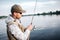 A picture of man in cap, sunglasses and vest standing in shallow and looking at fly rod. He is using non-inertial reel