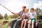 A picture of man and his children sitting together on the river shore. Guy is fishing while his kids are watching on it