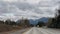The picture of the road with vehicles and mountains in front with clouds over them