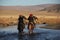 Picture of two horseriders in a river surrounded by a valley with hills on a blurry background