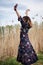 Picture of young blond woman, wearing dark boho dress dancing in front of dry yellow grass in field. Hippie musician relaxing in