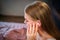 Picture of young woman sitting on bed. She holds hand close to ear. She points on it. It is closed with piece of cotton