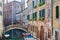A picturesque view of a venetian canal, with the colourful historic houses overlooking it, a pedestrian bridge and boats moored, o