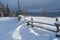 Picturesque waved shadows on snow from wood fence. Alpine mountain winter hamlet outskirts, snowy path, fir forest on far misty