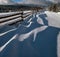 Picturesque waved shadows on snow from wood fence. Alpine mountain winter hamlet outskirts, snowy path, fir forest. High