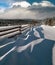 Picturesque waved shadows on snow from wood fence. Alpine mountain winter hamlet outskirts, snowy path, fir forest. High