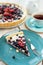 Piece of blueberry open pie on blue plate with cup of black tea and white tea pot on bright painted wood background