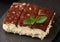 A piece of tiramisu sprinkled with cocoa, on top of a sprig of fresh mint on a black background