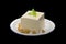 A piece of tofu served on a plate, decorated with green leaves, placed on a black background