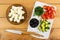 Pieces of cheese in saucer, bowls with dill, black olives, pieces of cucumbers, tomato, knife on cutting board on table. Top view