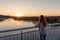 On a pier with steel railing over water stands girl young woman with her back with curly hair at sunset