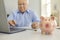 Piggy bank standing on desk with blurred senior man using laptop to pay bills online in background