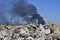 A pile of concrete rubble with protruding rebar on the background of thick black smoke in the blue sky.