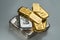 A pile of gold and silver bars of various weights from different manufacturers lies on a grey background