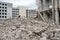 A pile of gray concrete fragments of the remains of a building against the background of a large destroyed building