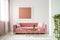 Pillows on pink sofa in white apartment interior with painting and flowers on copper table. Real photo