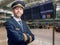 Pilot in uniform with golden stripes and cap crossed his arms while standing in the airport