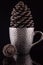 Pimpled silver coffee cup with a large pinecone inside on the reflective black surface. And the second small pinecone