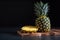 Pineapple stands on a wooden board on a dark background. On the left is a slice of pineapple