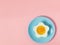 Pink background with egg in a flower shaped bowl and blue plate with broken egg shells and kitchen utensils