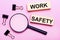 On a pink background, a magnifier, black paper clips and wooden blocks with the text WORK SAFETY. Business concept