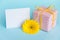 Pink dotted gift box, yellow gerbera flower and empty card over a blue background.
