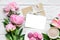 Pink peony flowers and blank greeting card with cappuccino cup, macaroons and gift box on white wooden table
