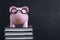 Pink piggy bank wearing eyeglass on top of stack of books with dark blackboard or chalkboard background using as cost of knowledge