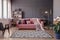 Pink sofa with two blankets and cushions standing in sitting room interior with windows with curtains, patterned carpet and empty