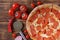 Pizza with salami, pepperoni and red chilli lies on a plank table next to tomatoes and a special circular knife