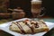 Plate of Crepe with Chocolate Sauce and Almond Nuts, with Blurred Hot Cappuccino and Sugar Pot in Background