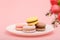 Plate of macaroons on pink background with flowers. Sweet pastry, baked products, sweets, dessert. Unhealthy diet